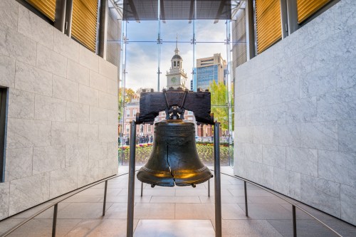 The Liberty Bell displayed indoors, framed by modern architecture, with a historic clock tower visible outside.