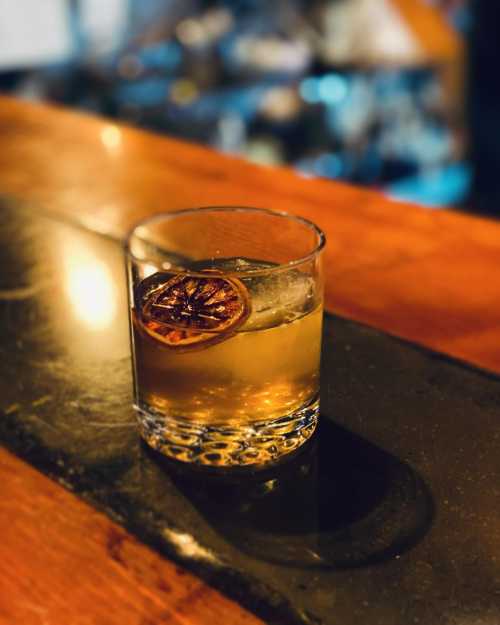 A glass of cocktail with ice and a dried citrus slice, resting on a dark stone surface. Blurred bar background.