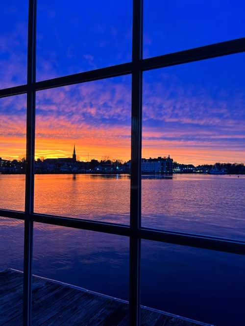 A vibrant sunset over a calm lake, viewed through a window with reflections and silhouettes of buildings.