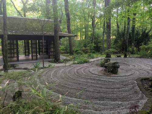 A serene garden with a raked gravel path, surrounded by trees and a wooden structure in the background.