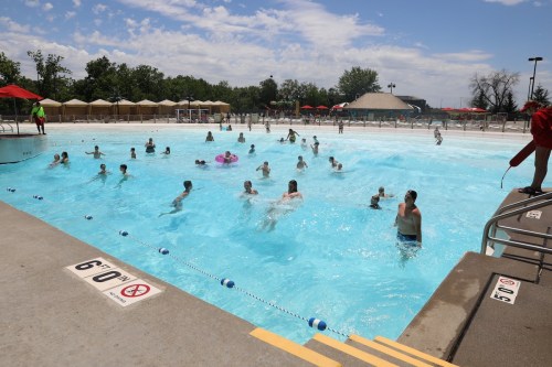 A busy outdoor swimming pool with children and adults enjoying the water on a sunny day. Red umbrellas provide shade.