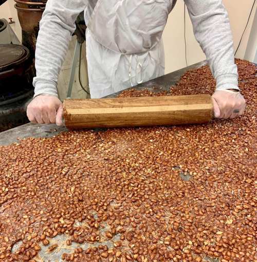 A person in an apron rolls a wooden pin over a large spread of roasted nuts on a table.