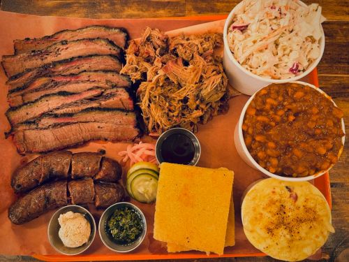 A vibrant platter featuring sliced brisket, pulled pork, sausage, coleslaw, baked beans, cornbread, and mashed potatoes.