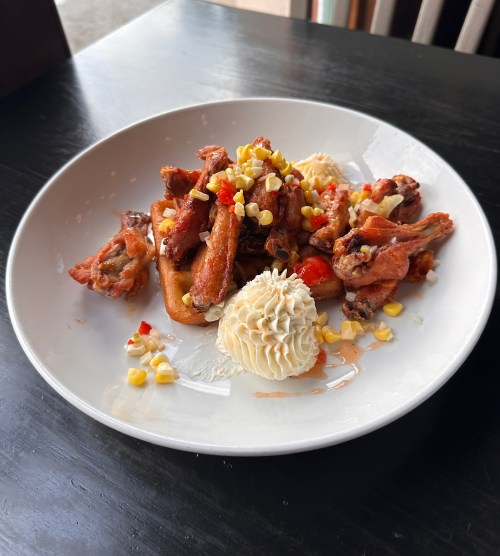 A plate of crispy chicken wings on a waffle, topped with corn, diced peppers, and a dollop of whipped cream.