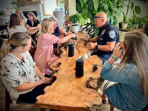 A police officer sits at a wooden table with several women, enjoying tea and conversation in a cozy setting.