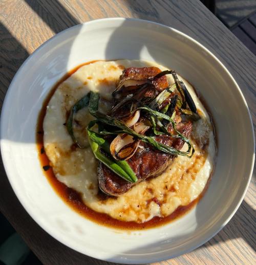 A plate of tender meat atop creamy grits, garnished with green onions and a rich sauce, served in natural light.