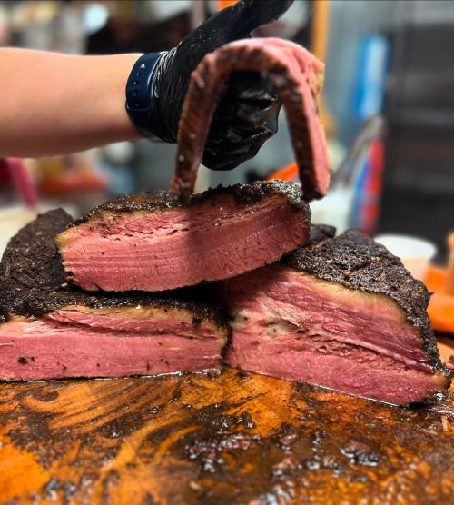A hand wearing a black glove holds a slice of smoked brisket, showcasing its juicy, pink interior on a wooden cutting board.