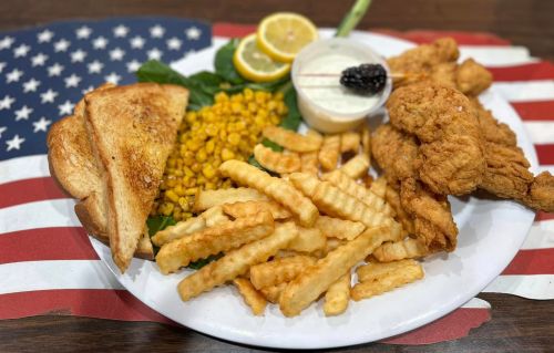 A plate of fried chicken, crinkle-cut fries, corn, toast, and a dipping sauce, set on a patriotic American flag background.