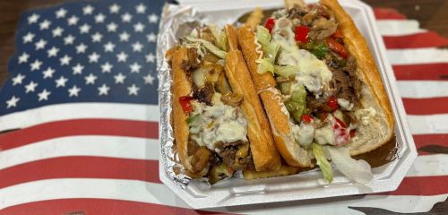 Two loaded sandwiches with meat, vegetables, and sauce, served in a takeout container on an American flag background.