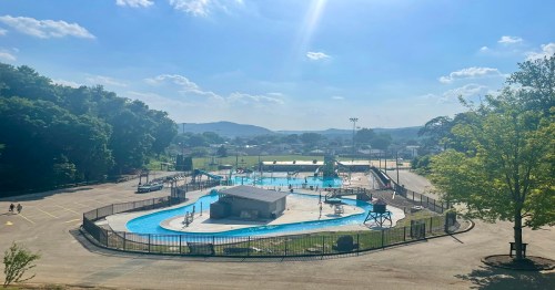 A sunny view of a water park with a pool, slides, and surrounding greenery, set against a blue sky and distant hills.