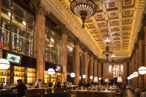 Elegant restaurant interior with high ceilings, ornate decor, and a long bar, featuring patrons dining and enjoying drinks.
