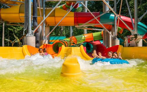 Two groups of people enjoy colorful water slides, splashing into a bright yellow pool below on a sunny day.