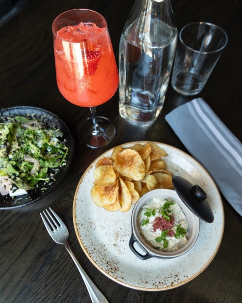 A plate with creamy dip, chips, a salad, a cocktail, and a bottle of water on a dark wooden table.