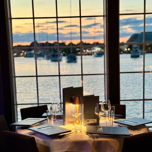 A cozy dining table by a window, overlooking a sunset on the water, with menus and candlelight.