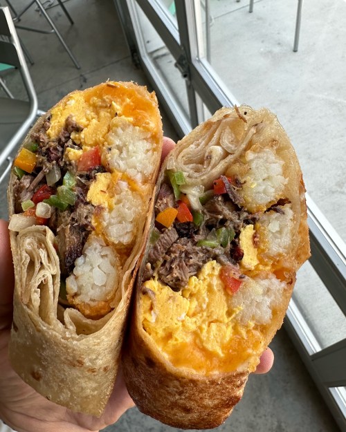 A hand holding a halved burrito filled with scrambled eggs, beef, and colorful vegetables, showcasing its hearty ingredients.