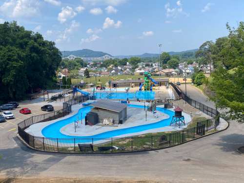 Aerial view of a water park featuring a blue pool, slides, and surrounding green hills under a clear sky.