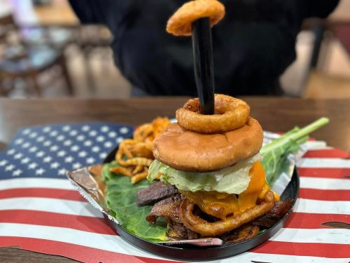 A towering burger with cheese, lettuce, and onion rings, served on a plate with curly fries and a patriotic backdrop.