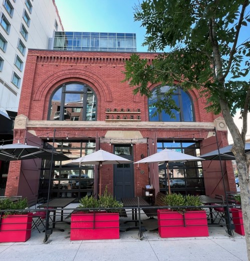 Exterior of a red brick restaurant with large windows, outdoor seating, and umbrellas, surrounded by greenery.