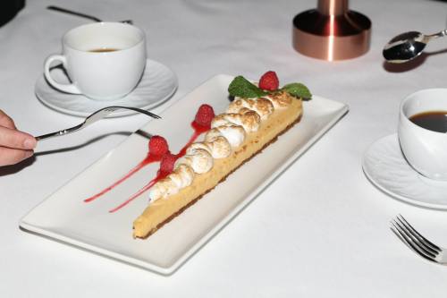 A slice of lemon meringue tart with raspberries and mint, served with coffee on a white tablecloth.