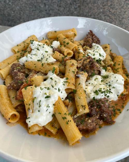 A bowl of rigatoni pasta topped with meat, creamy ricotta, and sprinkled with herbs.