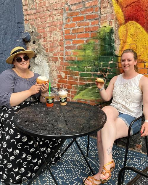 Two women sit at a table with drinks and pastries, smiling in front of a colorful brick wall.