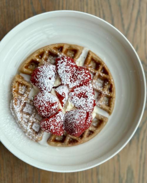A plate of waffles topped with fresh strawberries and powdered sugar, served in a white bowl on a wooden table.