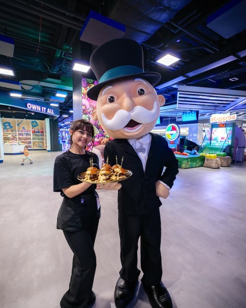A woman in a black shirt holds a tray of food next to a large mascot in a tuxedo and top hat.