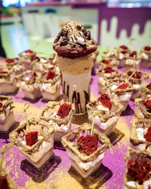 A colorful display of dessert cups topped with red velvet cake and a large milkshake with cookie crumbles.