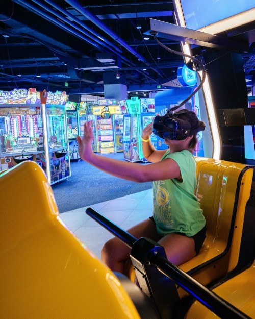A child wearing a VR headset sits in a yellow arcade game, reaching out with hands in a colorful gaming environment.