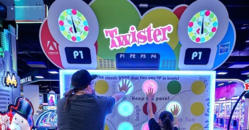 Two people play a giant Twister game at an arcade, reaching for colored circles on a vibrant game board.