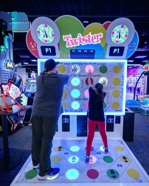 A man and a girl play a digital Twister game, reaching for colored circles on a bright arcade machine.