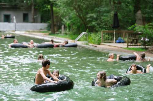 Children and adults float on inner tubes in a green-tinted pool surrounded by trees and a wooden deck.