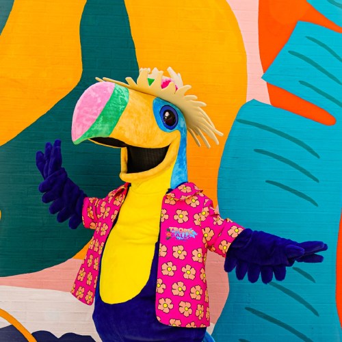A colorful parrot mascot wearing a floral shirt and a straw hat, posing in front of a vibrant mural.