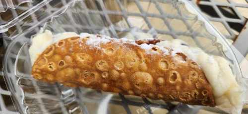 A close-up of a cannoli filled with cream, dusted with powdered sugar, in a clear plastic container.