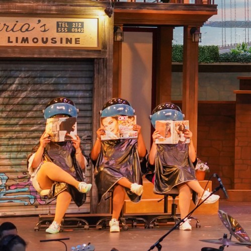 Three people in salon chairs with blue hair dryers, holding magazines, on a stage with a backdrop of a cityscape.