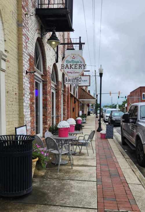 A quaint street scene featuring a bakery sign, outdoor seating, and planters along a brick sidewalk on a cloudy day.