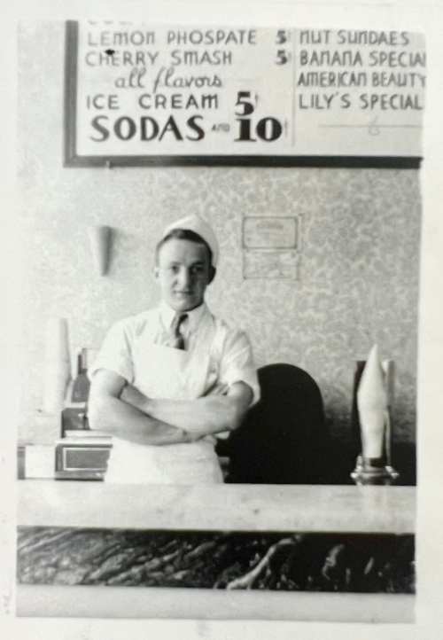 A vintage black-and-white photo of a young man in an ice cream shop, standing behind a counter with a menu above.
