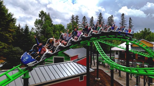 A roller coaster with excited riders going down a steep drop, surrounded by trees and colorful buildings.