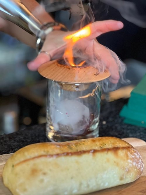 A hand holds a wooden lid over a glass with smoke and flames, next to a toasted bread roll on a wooden board.