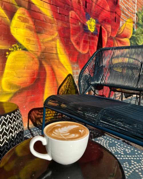 A white coffee cup with latte art sits on a table in front of a colorful flower mural and a blue chair.