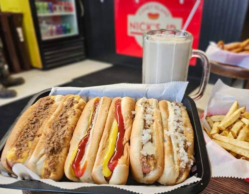 A tray with four hot dogs topped with various condiments, alongside a glass of milkshake and a side of fries.
