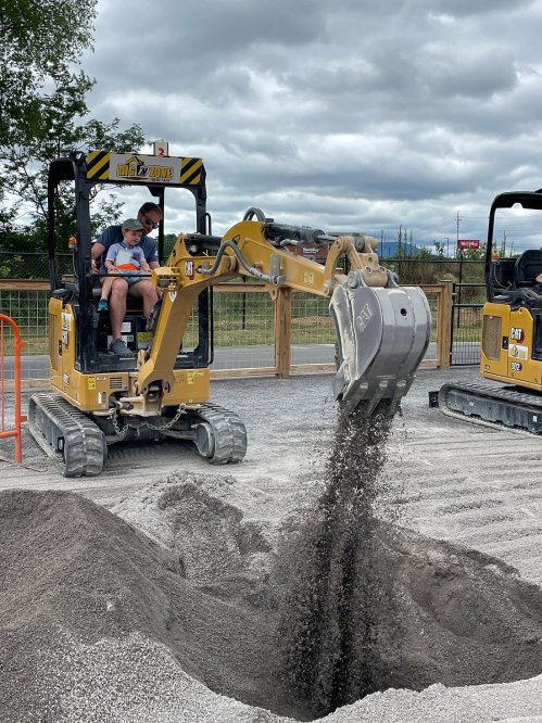 A child operates a small excavator, digging into a pile of gravel at a construction site under cloudy skies.