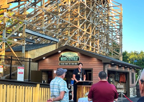 A speaker addresses an audience in front of a wooden structure, likely a roller coaster, at an outdoor event.