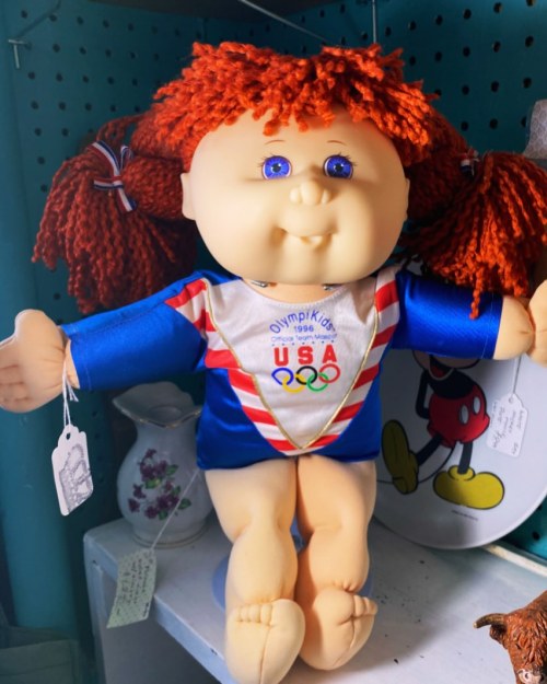 A vintage Cabbage Patch doll wearing a blue Olympic-themed outfit with red pigtails and blue eyes.