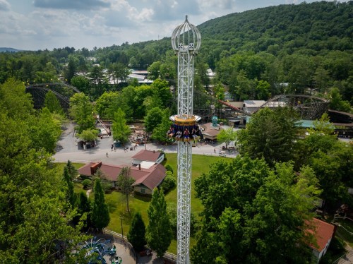 Aerial view of a theme park featuring a tall drop tower ride surrounded by trees and other attractions.