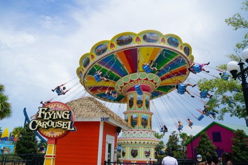 A colorful flying carousel with riders swinging in the air, surrounded by vibrant buildings and a clear blue sky.