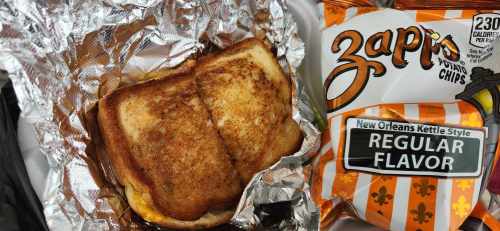 A grilled cheese sandwich wrapped in foil next to a bag of Zapp's regular flavor potato chips.