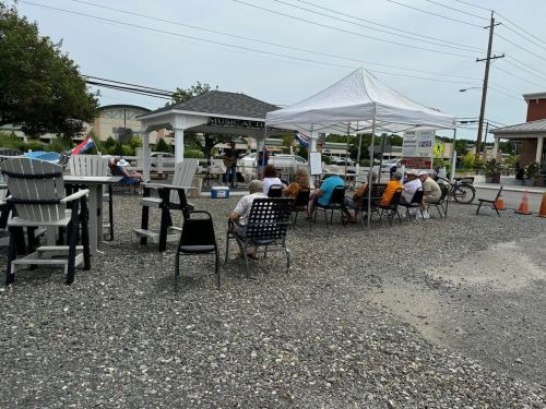 A small outdoor gathering with chairs set up under a tent, people seated, and a stage in the background.