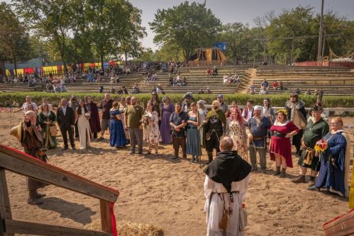 A group of people in medieval costumes gathers in a sandy arena, surrounded by trees and spectators in the background.