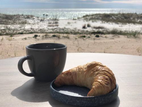 A croissant on a plate beside a coffee cup, with a scenic beach and ocean in the background.